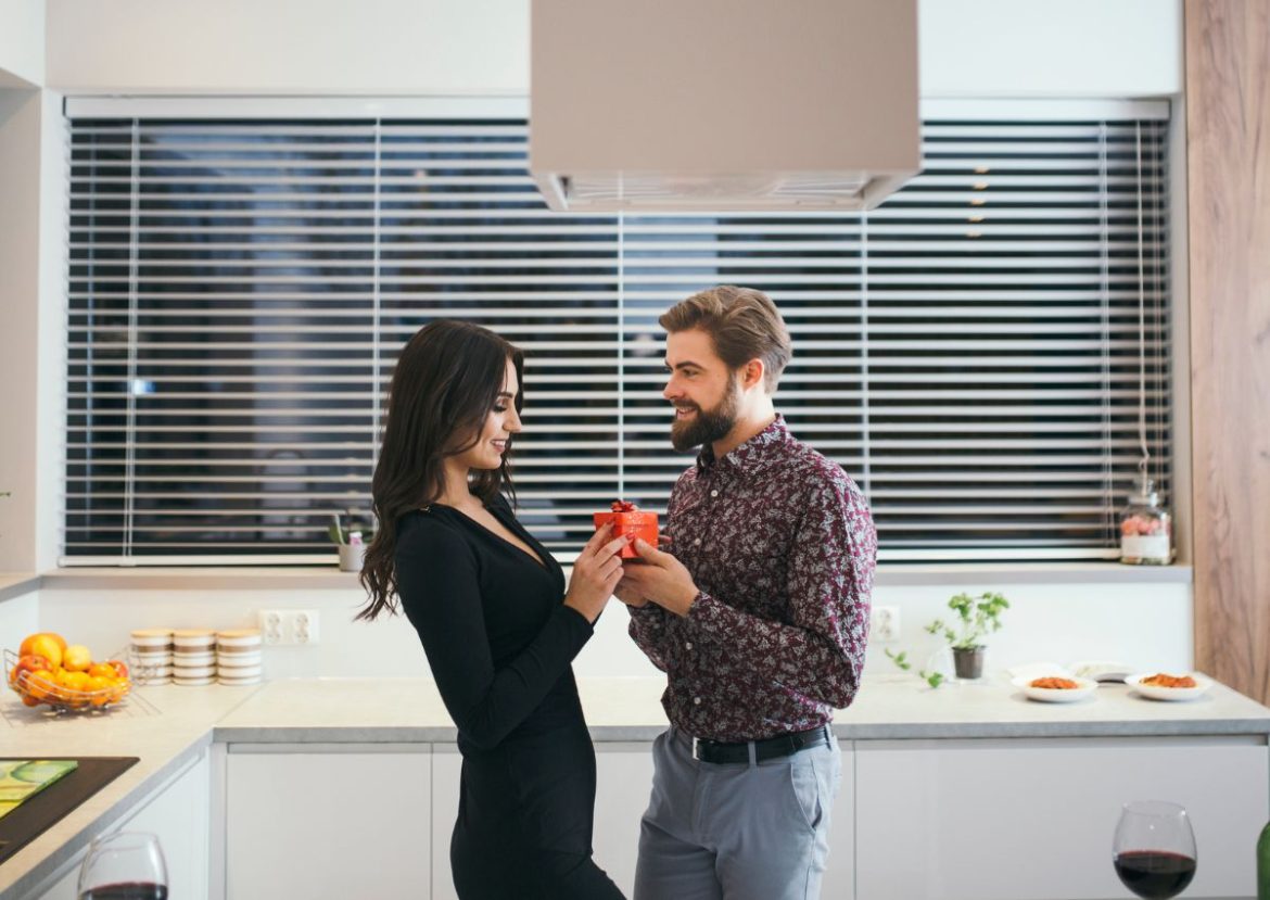 Un couple souriant échange un cadeau dans une cuisine moderne équipée de stores sur mesure.