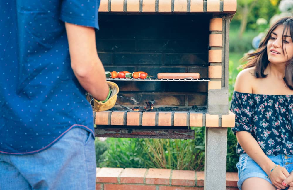 Un homme utilise une pince pour cuire des brochettes sur un barbecue en brique pendant qu'une femme le regarde.